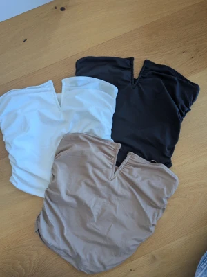 Tre ärmlösa tubtoppar med v-ringning - Säljer tre stilrena tubtoppar i vitt, svart och beige. Alla har en snygg v-ringad detalj framtill och är ärmlösa, perfekta att matcha med jeans eller kjol. Materialet känns mjukt och stretchigt, troligen polyester eller syntet. Magtröjar korta.