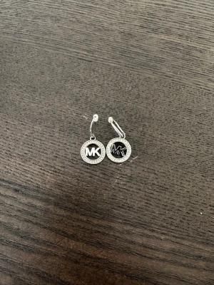 Silverörhängen från Michael Kors - Säljer ett par snygga silverörhängen från Michael Kors med rund form och MK-logga i mitten. Örhängena har gnistrande stenar runt kanten som ger en lyxig känsla. Perfekt för dig som gillar stilrena accessoarer med designer-touch.