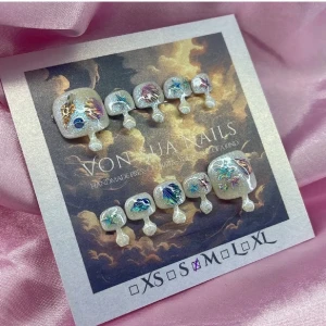 Shelly (M);Square Toes - Unika tå-press-on naglar i storlekM: 16 mm, 9,5 mm, 10 mm, 9,5 mm, 7 mm . Naglarna har en skimrande bas i cateyesilver och vitt med detaljer i blått, lila och samt inslag av glitter och holografiska effekter. Fyrkantig form och fantasifulla motiv gör dem till ett statement.