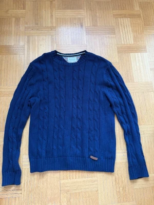 Mörkblå kabelstickad tröja Bondelid - Mörkblå kabelstickad tröja från Bondelid med rund halsringning och ribbade muddar. Klassisk design med diskret läderdetalj nertill. Perfekt för lager-på-lager och snygg till jeans eller chinos. Skriv vid funderingar ☺️