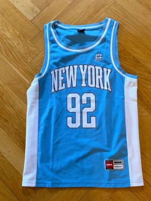 Blå New York basketlinne - Snyggt basketlinne i ljusblått med vita sidopaneler och tryck. Stort 'NEW YORK' och siffran 92 framtill. Klassisk sportig look med ribbade kanter och broderad patch nertill. Perfekt för träning eller streetstyle.