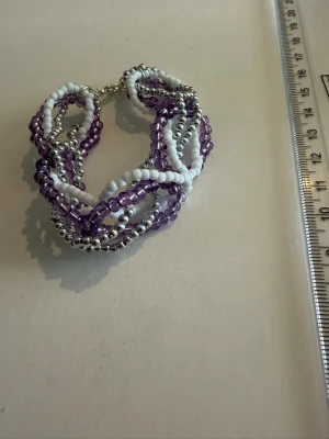 Pärlarmband i lila och vitt - Säljer ett snyggt armband med flera rader av små pärlor i lila, vitt och silver. Armbandet har en tvinnad design och stängs med ett metallspänne. Perfekt accessoar för att ge din outfit lite extra färg och personlighet.