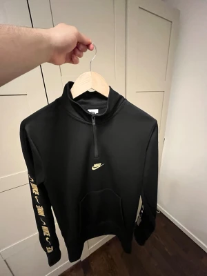 Svart Nike hoodie med dragkedja - Svart hoodie från Nike med halv dragkedja framtill och gul Nike-logga på bröstet. Ärmarna har flera Nike-loggor i gult längs sidan. Praktisk magficka och sportig look