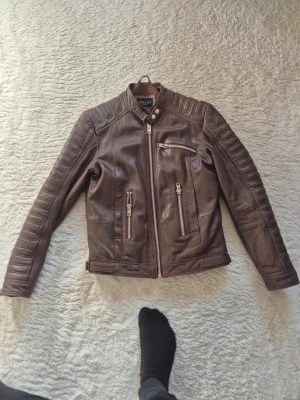 Brun skinnjacka från Hollies - Snygg brun skinnjacka från Hollies med coola detaljer och quiltade partier på axlar och rygg. Jackan har tre dragkedjeförsedda fickor framtill, ståkrage med tryckknapp och justerbara spännen i midjan. Perfekt för dig som gillar en tuff och stilren look.