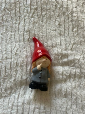 Keramiktomte med röd luva - Söt liten tomtefigur i keramik med röd spetsig luva, grå jacka och svarta skor. Tomten har långt blont hår och håller ett finger mot kinden. Perfekt som dekoration till jul eller att ställa på hyllan.