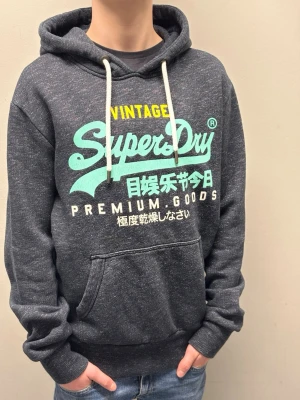 Vintage Superdry hoodie - Riktigt fin vintage superdry hoodie men nån blandning av mörkblå svart och grå färg med vita inslag i tyget. Riktigt fint tryck som är typ som spräcklig färg eller vad man ska kalla det. Riktigt fint skick i stl s. Inga slitningar eller andra tecken på användning
