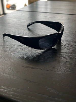 Svarta solglasögon från Prada - Snygga svarta solglasögon från Prada med sportig, bred båge och mörka glas. Klassisk logga på sidan och en clean, futuristisk look som passar perfekt till streetwear eller en stilren outfit. Tillverkade i blank syntet för en modern vibe.