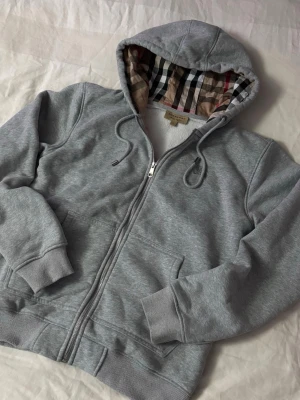 Grå zip-hoodie från Burberry - Snygg grå hoodie med dragkedja från Burberry. Klassisk huva med det ikoniska rutmönstret på insidan, dragsko och två fickor framtill. Mjuk bomullsblandning och ribbade muddar vid ärmar och midja. Perfekt för en chill och stilren look.