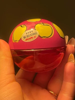 DKNY Be Delicious Orchard St parfym 30ml - Säljer en DKNY Be Delicious Orchard St parfym i en rund, halvt genomskinlig röd flaska med rosa lock dekorerat med gula äpplen. Flaskan har en lekfull och färgstark design som sticker ut i badrumshyllan.