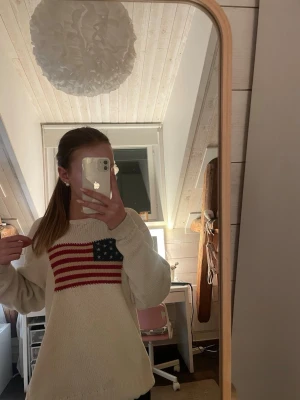 Stickad tröja med amerikansk flagga - Stickad tröja i offwhite med ett stort motiv av den amerikanska flaggan på bröstet. Materialet är mjukt och grovt stickat, perfekt för att skapa en avslappnad look. Det är riktig autentisk Ralph lauren. Storleken vet jag inte helt det står XL men är mer som storlek M💕