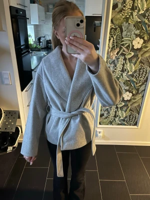 Grå omlottkappa med knytskärp - Säljer en stilren grå omlottkappa med bred krage och knytskärp i midjan. Kappan har en mjuk och lite tjockare känsla, perfekt för kyliga dagar. Modellen är rak med långa ärmar och passar dig som gillar minimalistisk design. Säljer på grund av att den inte kommer till användning 