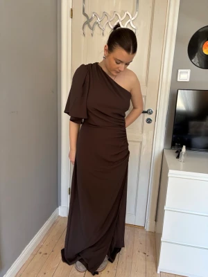 Brun oneshoulder balklänning - Elegant brun långklänning med oneshoulder-design och kort ärm på ena sidan. Klänningen har draperad midja och faller snyggt längs kroppen. Perfekt för dig som vill ha en stilren och modern look.