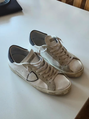 Philippe Model vita sneakers med svart detalj - Snygga vita sneakers från Philippe Model med beige mockadetaljer och svart hälparti. Klassisk låg modell med vita snören och ikonisk sköldlogga på sidan. Ovandel i läder och mocka, perfekt nu till våren.