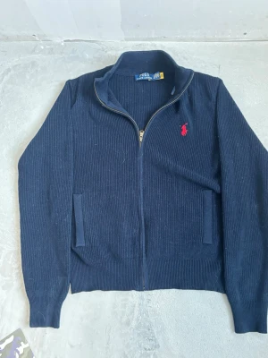 Mörkblå zip-tröja från Polo Ralph Lauren - Mörkblå stickad zip-tröja från Polo Ralph Lauren med röd broderad logotyp på bröstet. Tröjan har hög krage, två fickor framtill och ribbade muddar vid ärmslut och nederkant. Perfekt för lager-på-lager och snygg till jeans.