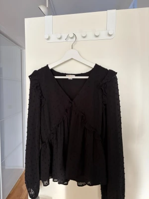 Svart volangblus med prickig struktur - Still Svart blus från Gina Tricot med volangdetaljer och diskret prickig struktur i tyget. Djup V-ringning och långa ärmar i transparent material. Köpt på Gina Young i strl 170 men har i vanliga fall Xs/S