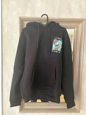 Svart hoodie från Casablanca - Svart hoodie från Casablanca med stor färgglad print på ryggen och mindre tryck på bröstet. Klassisk känguruficka framtill, dragsko i huvan och ribbade muddar. Perfekt för dig som gillar streetwear och snygga detaljer.