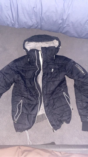 Svart pufferjacka med huva - Svart pufferjacka med huva och vita dragkedjor. Jackan har två snedställda fickor framtill, ribbade muddar och en ärmficka med dragkedja. Insidan är ljusgrå och jackan har en sportig look med diskret logga på bröstet.