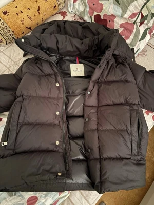 Svart dunjacka från Moncler - Snygg svart dunjacka från Moncler med hög krage och stora, quiltade partier. Jackan har dragkedja och tryckknappar framtill samt två stora fickor. Perfekt och riktigt trendig stil.