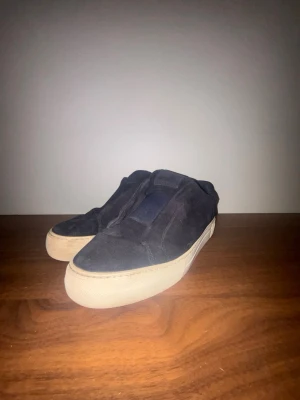 Svarta slip-on sneakers med vit sula - Säljer ett par svarta slip-on sneakers med vit, tjock gummisula. Skorna har en enkel och clean design utan snörning och är tillverkade i tygmaterial. Perfekta för dig som gillar minimalistisk stil och vill ha något lätt att ta på sig.