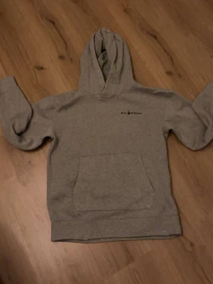 Grå sail racing hoodie med magficka - En enkel och stilren grå hoodie med huva och stor magficka framtill. Liten diskret logga på bröstet. Tillverkad i mjukt sweatshirtmaterial, perfekt för en avslappnad look. Långa ärmar och ribbade muddar vid ärmslut och nederkant.