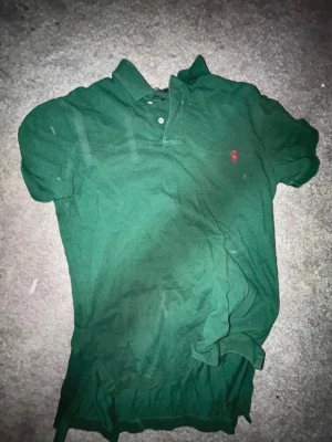 Grön pikétröja från Ralph Lauren - Klassisk grön pikétröja från Ralph Lauren med korta ärmar och krage. Tröjan har två knappar vid halsen och den ikoniska rosa logotypen broderad på bröstet. Tillverkad i mjuk bomull för en bekväm känsla.