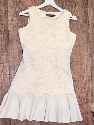 Vit kortklänning från Vita Rose - Supersnygg vit kortklänning från Vita Rose med ärmlös design och subtilt mönster framtill. Klänningen har en figurnära passform och volang längst ner för extra stilpoäng. Perfekt för dig som vill sticka ut med en elegant look.