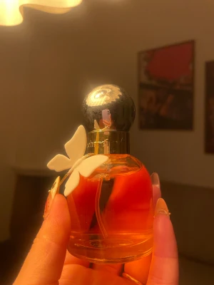 Cacharel Ella Ella 50ml - Säljer en elegant parfym från Cacharel, i en rund glasflaska med guldfärgad vätska och dekorativ vit duva på flaskhalsen. Svart, glansig kork med blommönster. Perfekt för dig som gillar klassiska dofter och snygg design.