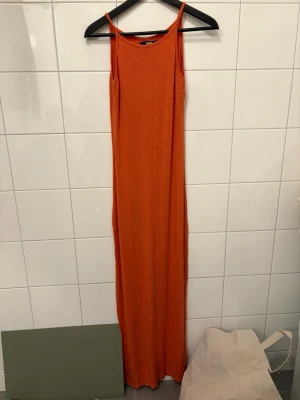 Orange långklänning från Bik Bok - Snygg orange långklänning från Bik Bok med smala axelband och rak passform. Klänningen är ärmlös och tillverkad i ett mjukt bomullsmaterial, perfekt för varma dagar. Enkel och stilren design som är lätt att matcha med accessoarer.
