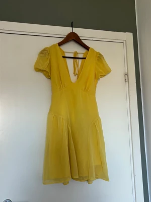 Gul kort klänning  - Söt gul klänning med korta ärmar och djup v-ringning både fram och bak. Klänningen har knytband i ryggen och är gjord i ett lätt, tunt tyg 