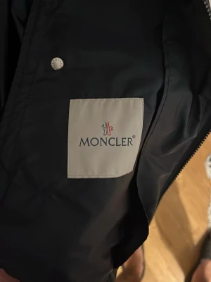 Navy Moncler vindjacka med huva - Snygg navy vindjacka från Moncler med huva och dragkedja. Jackan har diskreta röda och vita detaljer på axlarna och klassisk Moncler-logga på insidan. Tillverkad i ett lätt och glansigt material som skyddar mot vind. Perfekt för en stilren och sportig look.
