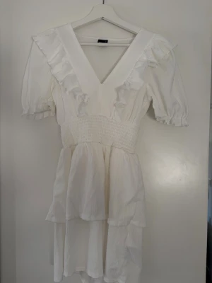 Vit volangklänning från Bik Bok - Superfin vit klänning från Bik Bok med v-ringning, volangdetaljer och korta puffärmar. Klänningen har smock i midjan och är tillverkad i ett lätt, strukturerat tyg som ger en luftig känsla. Perfekt för varma dagar och somriga tillfällen.