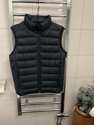 Mörkblå dunväst från Tommy Hilfiger - Snygg mörkblå dunväst från Tommy Hilfiger med diskret logga på bröstet och flagga i nacken. Västen har hög krage, två dragkedjeförsedda fickor och quiltad design. Perfekt för lager-på-lager och håller dig varm med stil. Skicket är 8/10. Det enda negativa är att en utav remarna i nacken har släppt men det är enkelt och fixa. Annnars har den fint skick. Storleken är 176 men passar som       xs/s också. Priset är ej hugget i sten. Hör av dig om du har frågor!