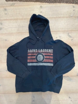 Mörkblå hoodie från Saint Laurent - Snygg mörkblå hoodie från Saint Laurent med tryck i rött, vitt och blått framtill. Den är helt ny med prislapp fast prislappen ligger bredvid. Klassisk huva med dragsko och känguruficka. Materialet är mjukt och skönt, perfekt för en avslappnad stil. Stor logga och grafiskt mönster på bröstet.