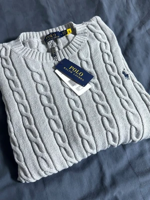 Ljusgrå kabelstickad tröja Polo Ralph Lauren - Kabelstickad tröja i ljusgrått från Polo Ralph Lauren med klassisk rund halsringning och mörkblå broderad logga på bröstet. Tröjan har ribbade muddar och är tillverkad i mjukt material som passar perfekt till lager på lager.