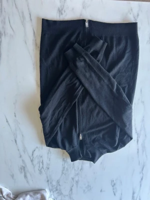 Svart tunn zip-kofta - Svart tunn kofta med dragkedja framtill och långa ärmar. Materialet är merinoull, perfekt för lager-på-lager. Enkel och stilren design utan synliga detaljer eller mönster. Hör av er vid fler frågor.