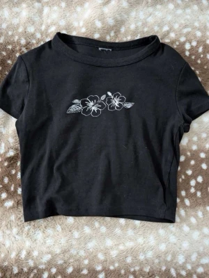 Svart croppad t-shirt med blommor - Svart croppad t-shirt med vit broderad blomdetalj framtill. Kortärmad och rund halsringning, perfekt för en trendig och avslappnad stil.