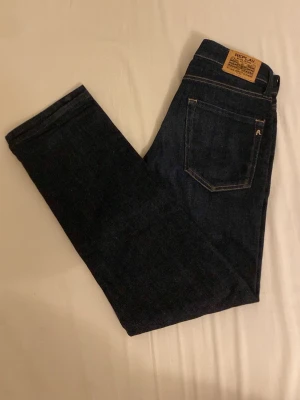 Mörkblå jeans från Replay - Mörkblå jeans från Replay med klassisk femficksdesign, kontrastsömmar och läderpatch med logga bak i midjan. Jeansen är i denim och har en stilren look med raka ben och metallogo på bakfickan. Pristet är diskuterbart.