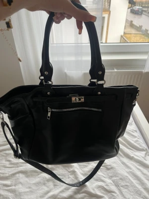 Svart handväska med axelrem - Snygg svart handväska i slätt konstläder med både handtag och avtagbar axelrem. Väskan har silverfärgade detaljer, dragkedjeficka framtill och ett stilrent spänne. Perfekt för dig som vill ha plats för allt du behöver och gillar en klassisk look.