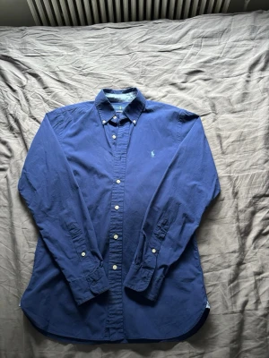 Marinblå skjorta från Ralph Lauren - Klassisk marinblå skjorta från Ralph Lauren i slim fit med button-down krage och vita knappar. Tillverkad i 100% bomull med stretch och har den ikoniska broderade loggan på bröstet. Perfekt för dig som gillar stilrena och tidlösa plagg.