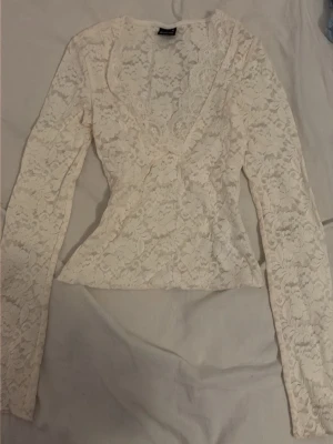 Vit spetsblus med v-ringning - Snygg vit/krämcvit blus i spets med v-ringning och långa ärmar. Blusen har en feminin och romantisk look med genombrutet spetsmönster och figurnära passform. Perfekt att styla med jeans eller kjol för en trendig outfit.