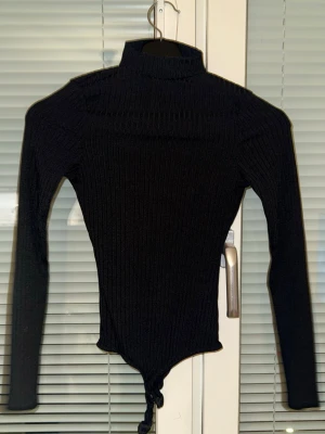 Svart ribbad polotopp med dragkedja - Svart ribbad polotopp med lång ärm och snygg half zip-dragkedja framtill. Toppen har en croppad passform med knytning nedtill och kan bäras både med hög krage eller nedvikt. Perfekt för en trendig och stilren look.