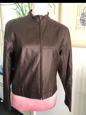 Brun minimalistisk jacka med dragkedja - Snygg brun jacka i minimalistisk stil med hög krage och tillverkad i ett lätt, det ser ut som bomullsmaterial. Jackan har hel dragkedja framtill, raka ärmar och två sidofickor. Passar perfekt för dig som <3 stilrena plagg.