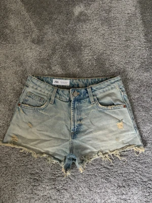 Ljusa jeansshorts från Zara - Säljer ett par ljusblå jeansshorts från Zara med fransig kant och slitna detaljer. Klassisk femficksmodell med knapp och dragkedja framtill.❣️