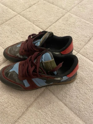 Valentino Garavani camo sneakers - Unika sneakers från Valentino Garavani med camomönster i blått, svart och grönt. Skorna har vinröda skosnören, röd häl och detaljer i mocka och läder. Låg modell med svart innersula och olivgrön plös med logga. Perfekta för dig som vill sticka ut.
