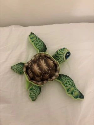 Grön plyschsköldpadda mjukisdjur - Söt mjukissköldpadda i grönt plyschmaterial med detaljerad sköld i brunt och grönt. Sköldpaddan har stora blå ögon och realistiskt mönstrade ben och huvud. Perfekt som prydnad eller gosedjur.