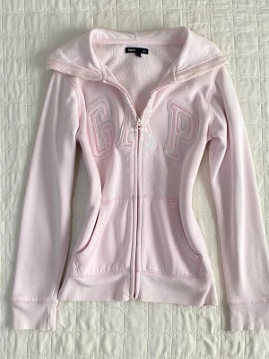 Ljusrosa hoodie från GAP - Snygg ljusrosa hoodie från GAP med dragkedja framtill och broderad logga på bröstet. Hoodien har två fickor, ribbade muddar och en klassisk huva. Perfekt för en avslappnad och trendig stil.