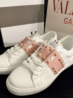 Valentino Garavani sneakers med nitar - Snygga vita sneakers från Valentino Garavani med rosa band över vristen dekorerat med ikoniska pyramidformade nitar. Skorna har klassisk låg modell, rund tå och vita snören. Tillverkade i läder för en lyxig känsla och stilren look.