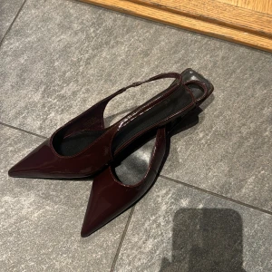 Vinröda lackade slingback-pumps - Säljer ett par vinröda slingback-pumps med spetsig tå och lackad finish. Skorna har öppen häl och smal rem runt ankeln. Skorna är helt oanvända stl 38