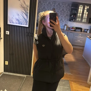 Svart dunväst från Adidas - Svart dunväst från Adidas med dragkedja och två fickor med knappar. Västen har en hög krage och Adidas-logga på bröstet. Perfekt för lager-på-lager och har en snygg, sportig look.