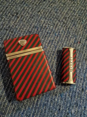 Gucci cigarettetui och tändarhållare - Säljer ett exklusivt accessoarset från Gucci med klassiska röd-gröna diagonala ränder och guldfärgade metalldetaljer. Setet innehåller ett cigarettefodral/etui och en tillhörande tändarfodrall, båda med tydlig Gucci-logga. Perfekt för dig som gillar lyxiga detaljer.
